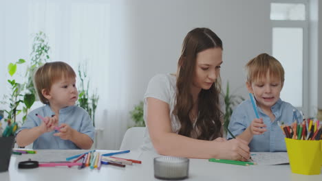 A-family-of-two-children-and-a-young-mother-sitting-at-the-table-draws-on-paper-with-colored-pencils.-Development-of-creativity-in-children.-white-clean-interior